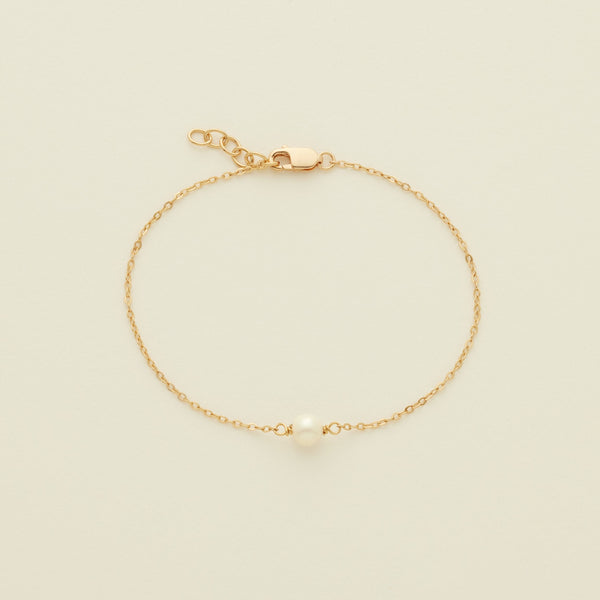 A delicate gold chain bracelet with a single white pearl at the center, displayed on a light cream background.
