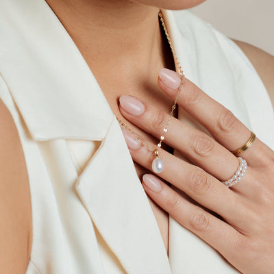 A close-up image of a person's hand resting on their chest, holding a dainty textured gold chain with a single pearl charm attached, paired with two stacked pearl strand rings and three stacked thin gold band rings.