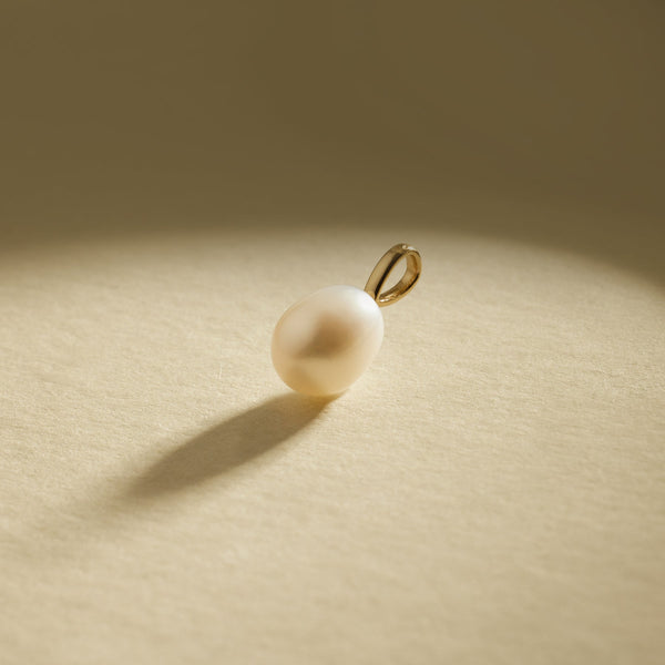 A pearl charm with a gold bail, set on a textured beige background with natural shadows.