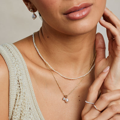 A close-up lifestyle image of a person wearing a pearl strand choker necklace paired with a textured gold chain necklace holding a square clear gemstone charm and a pearl charm.