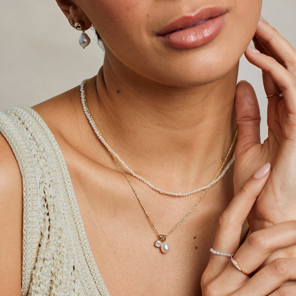 A close-up lifestyle image of a person wearing a pearl strand choker necklace paired with a textured gold chain necklace holding a square clear gemstone charm and a pearl charm.