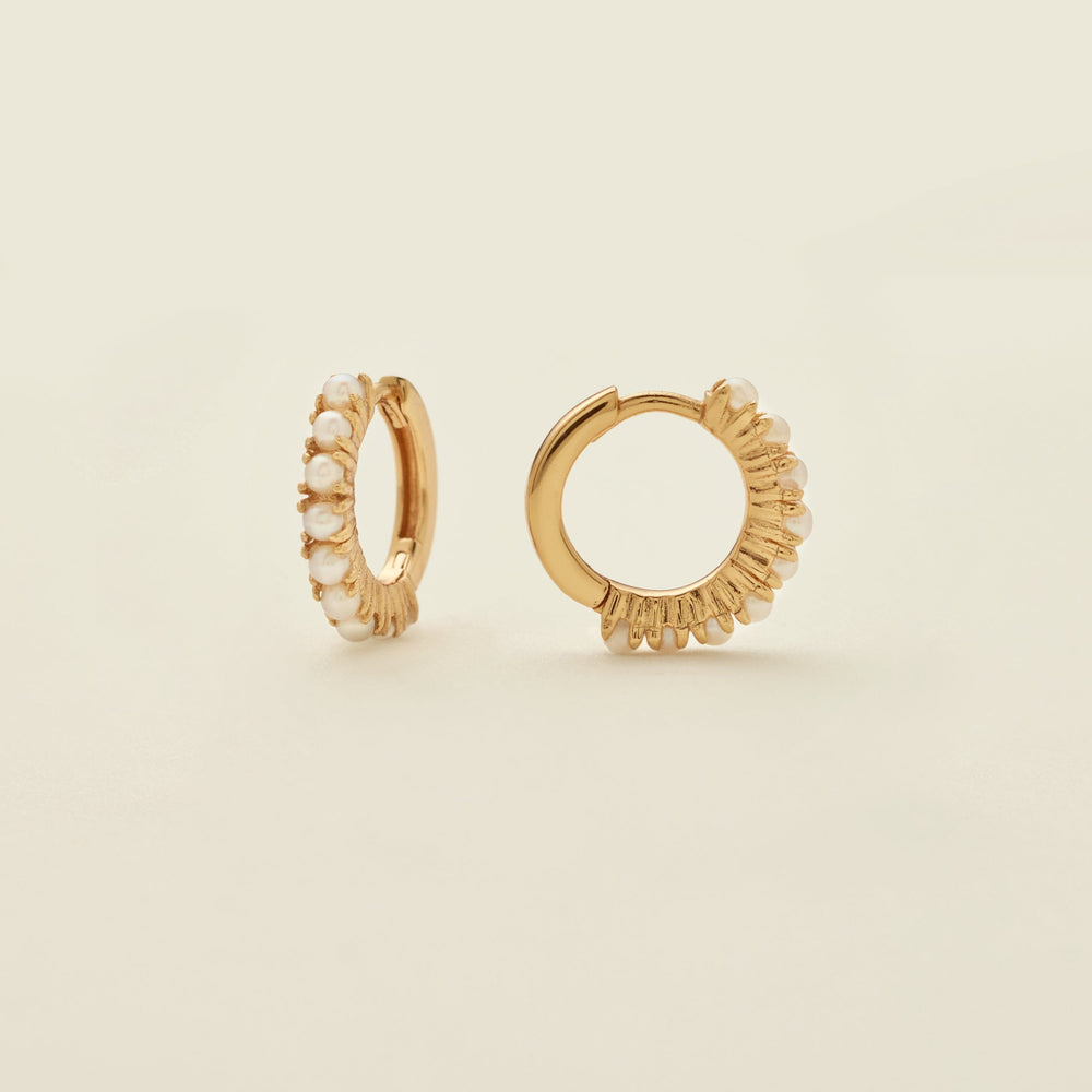 A pair of gold huggie hoop earrings adorned with small white pearls, shown on a clean, solid, light beige background.
