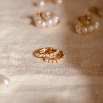 A pair of gold huggie hoops adorned with cream colored pearls laying on top of one another, shown on a light beige linen surface with other pieces of gold and pearl jewelry blurred in the background.