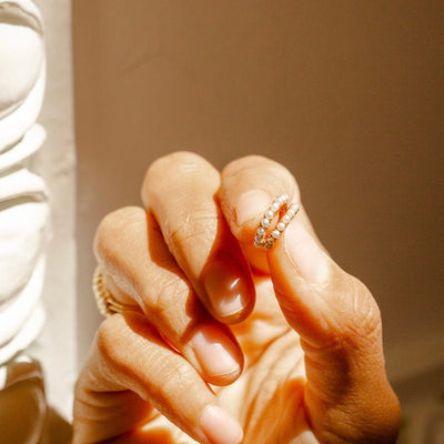 A person's fingers holding up a pair of gold huggie hoops adorned with round cream colored pearls in sunlight.