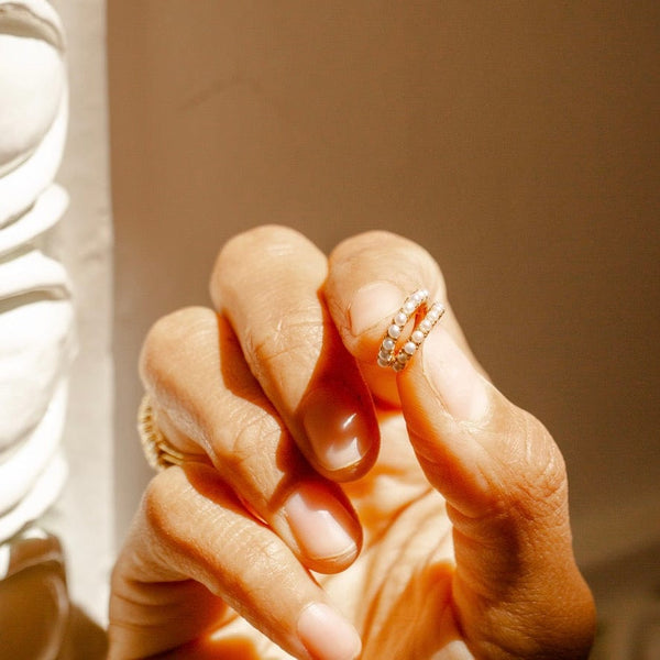 A person's fingers holding up a pair of gold huggie hoops adorned with round cream colored pearls in sunlight.