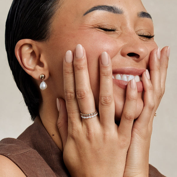 A close-up lifestyle image of a woman holding her hands to her face, showcasing a pearl strand ring and gold eternity ring with square light blue gemstones, paired with a gold stud earring with a peal hanging from it and a brown top.