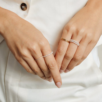 A close-up image of a persons hands wearing three pearl strand rings, three stacked thin gold band rings, and a gold ring with small clear gemstones across it.