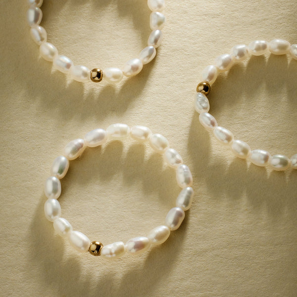 Three pearl strand rings, each with one gold bead, set against a textured beige background with natural shadows.
