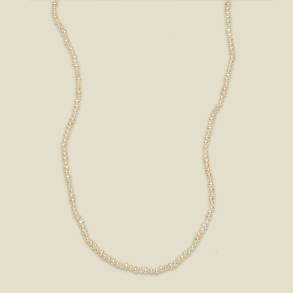 A delicate strand necklace made of small, irregular cream-colored pearls, arranged in a curved U-shape against a solid, warm off-white background.