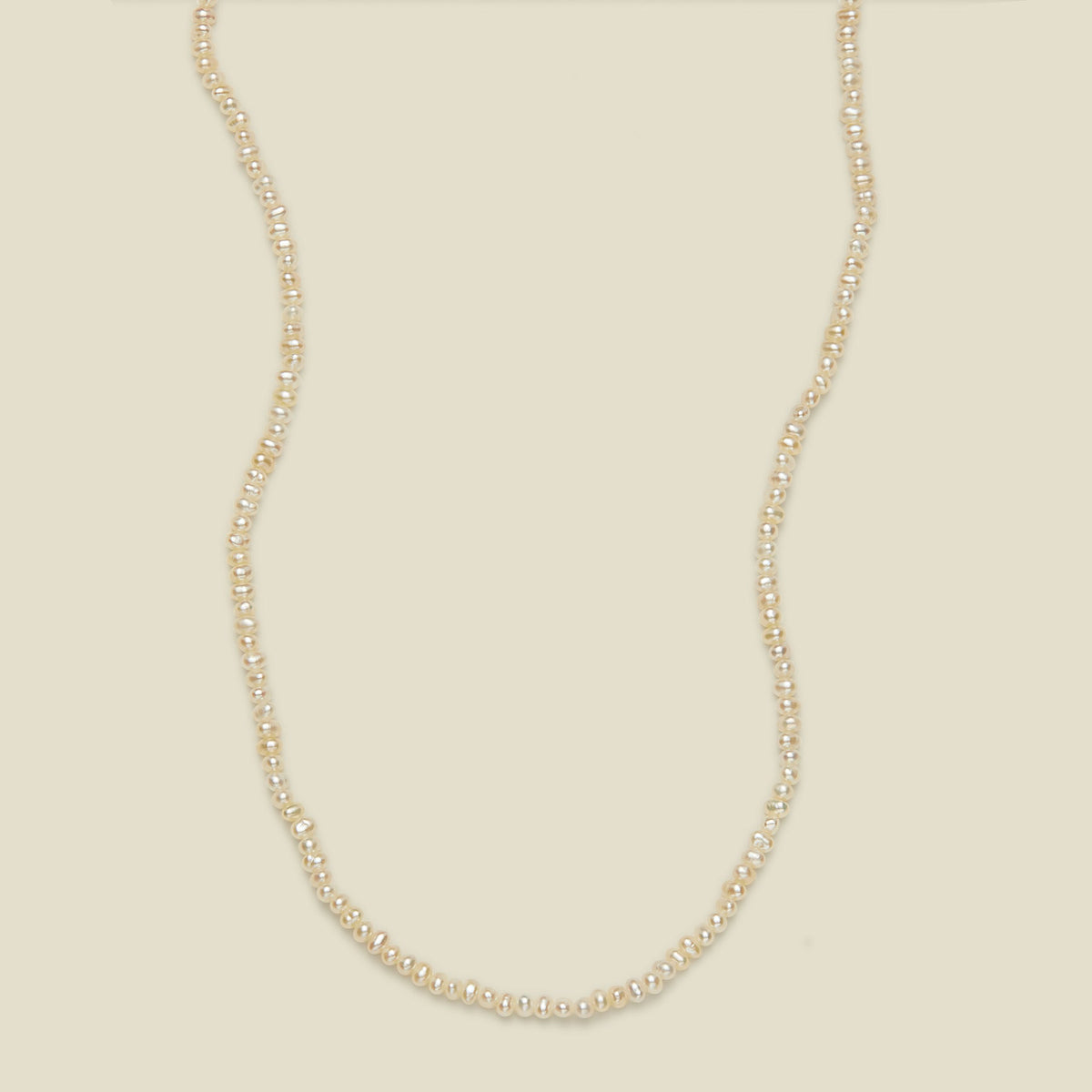 A delicate strand necklace made of small, irregular cream-colored pearls, arranged in a curved U-shape against a solid, warm off-white background.