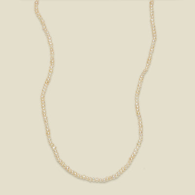 A delicate strand necklace made of small, irregular cream-colored pearls, arranged in a curved U-shape against a solid, warm off-white background.