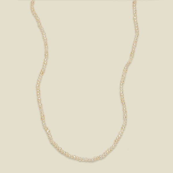 A delicate strand necklace made of small, irregular cream-colored pearls, arranged in a curved U-shape against a solid, warm off-white background.