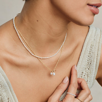 A close-up lifestyle image of a person wearing a pearl strand choker necklace paired with a textured gold chain necklace holding a square clear gemstone charm and a pearl charm.