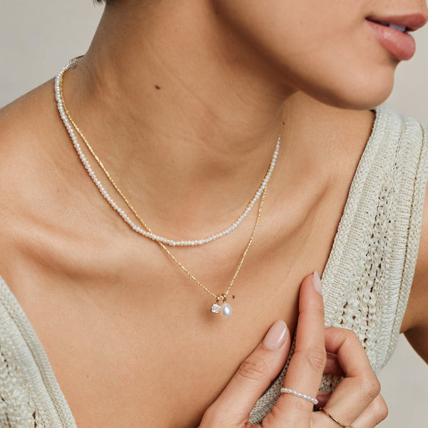 A close-up lifestyle image of a person wearing a pearl strand choker necklace paired with a textured gold chain necklace holding a square clear gemstone charm and a pearl charm.