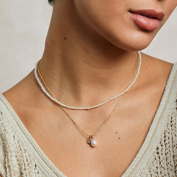 A close-up lifestyle image of a person wearing a pearl strand choker necklace paired with a textured gold chain necklace holding a square clear gemstone charm and a pearl charm.