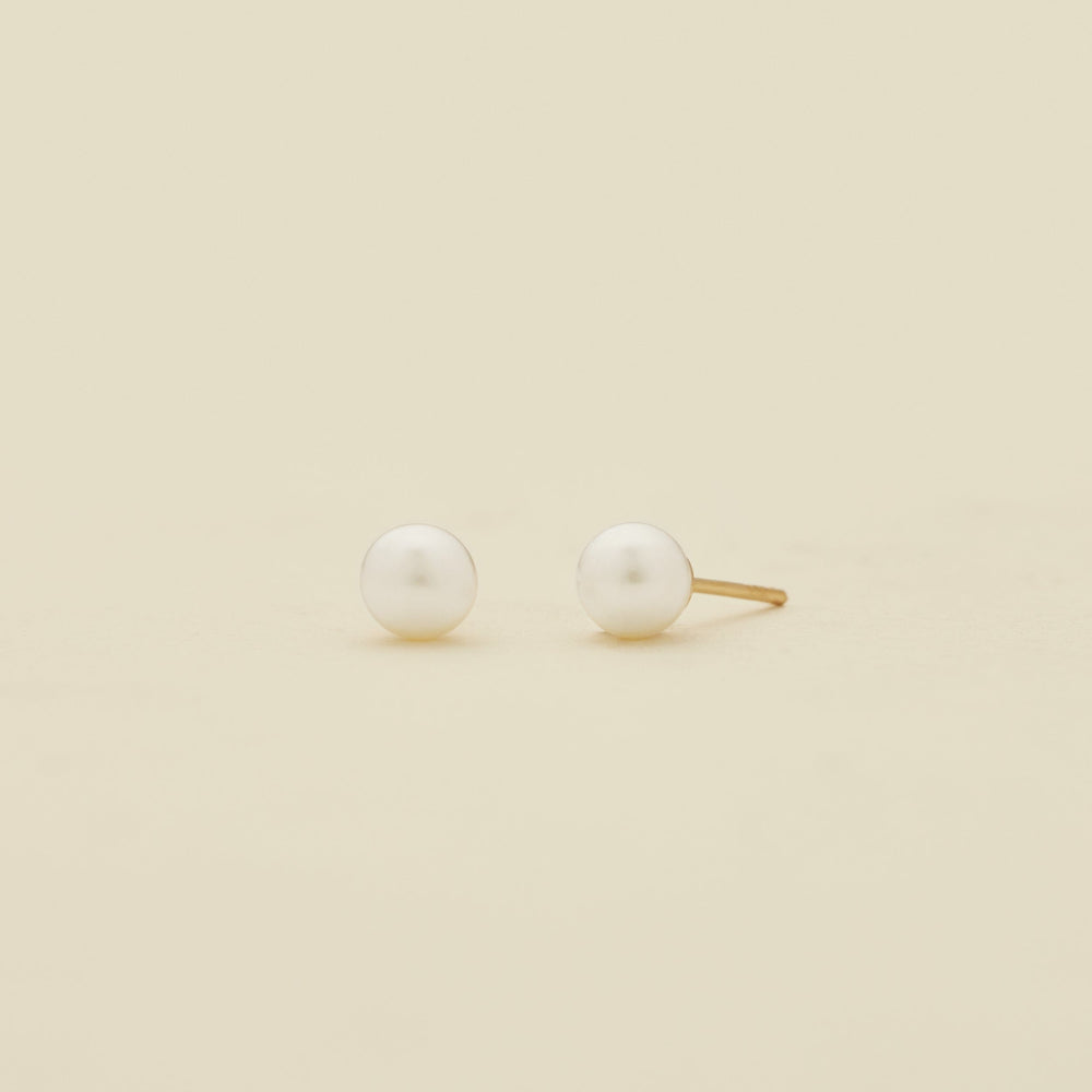 A pair of minimalist stud earrings featuring white round pearls set in a gold-tone post, displayed on a clean, solid cream-colored background.