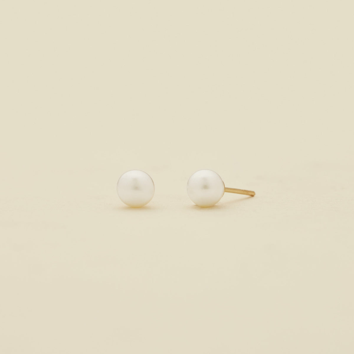 A pair of minimalist stud earrings featuring white round pearls set in a gold-tone post, displayed on a clean, solid cream-colored background.