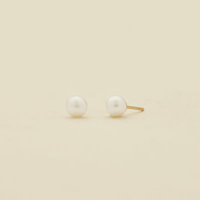 A pair of minimalist stud earrings featuring white round pearls set in a gold-tone post, displayed on a clean, solid cream-colored background.