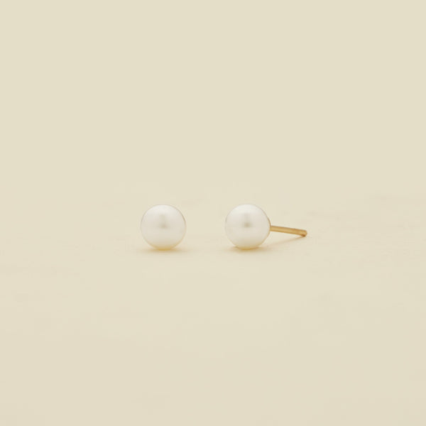 A pair of minimalist stud earrings featuring white round pearls set in a gold-tone post, displayed on a clean, solid cream-colored background.