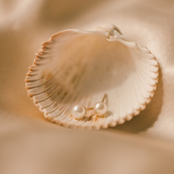 A pair of minimalistic round pearl stud earrings with gold posts, laying in a sea shell on a beige satin fabric.