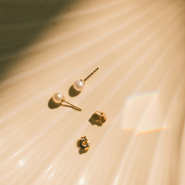 A pair of minimalistic round pearl stud earrings with gold posts laying next to two gold butterfly backings on a textured cream colored surface.