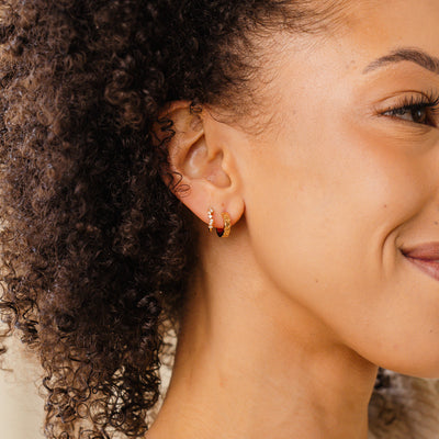 A close-up of a woman's ear wearing two gold hoops, one with clear round gemstones in a curving pattern and the other with the word 'PISCES' along the front in raised letters.