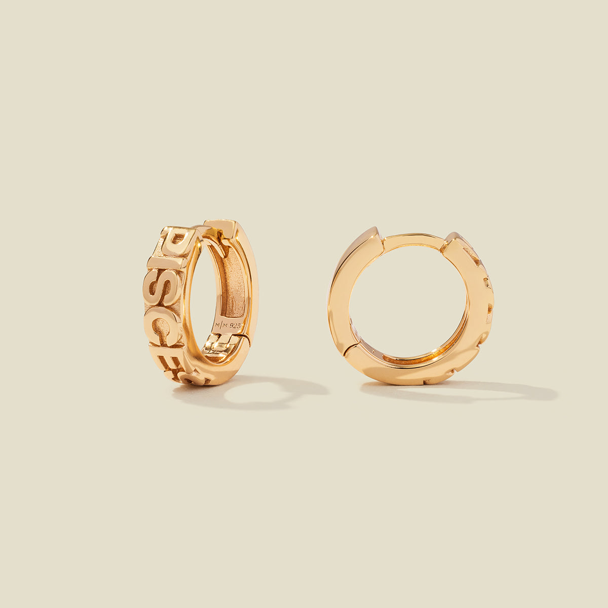 A pair of gold huggie hoop earrings, both standing upright, featuring raised lettering that spells 'PISCES' on the front. The earrings are photographed on a solid, warm beige background with soft shadows.