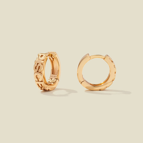 A pair of gold huggie hoop earrings, both standing upright, featuring raised lettering that spells 'PISCES' on the front. The earrings are photographed on a solid, warm beige background with soft shadows.