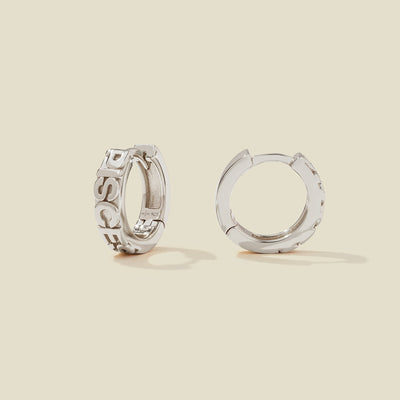 A pair of silver huggie hoop earrings, both standing upright, featuring raised lettering that spells 'PISCES' on the front. The earrings are photographed on a solid, warm beige background with soft shadows.