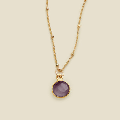 A gold chain pendant necklace with stationary gold beads, with a dark purple round gemstone charm, set against a solid beige background.