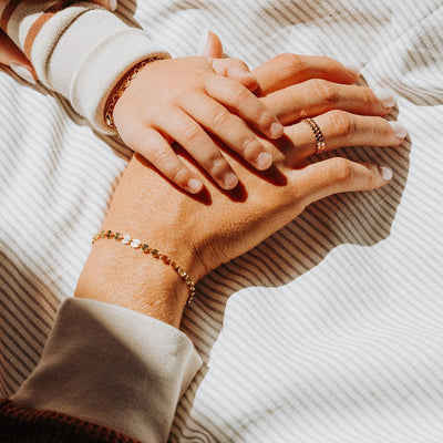 A close-up of a person's hand with a small child's hand resting on theirs, showcasing a gold chain bracelet with round flat linked discs and matching rings.