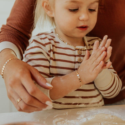 A young child with a mother's arm around her as they bake something in the kitchen, showcasing their matching gold chain bracelets with flat round linked discs.