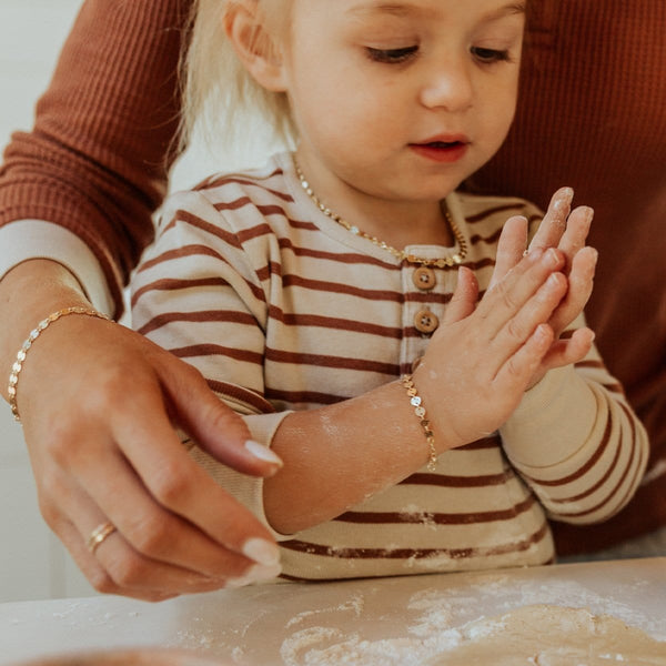 A young child with a mother's arm around her as they bake something in the kitchen, showcasing their matching gold chain bracelets with flat round linked discs.