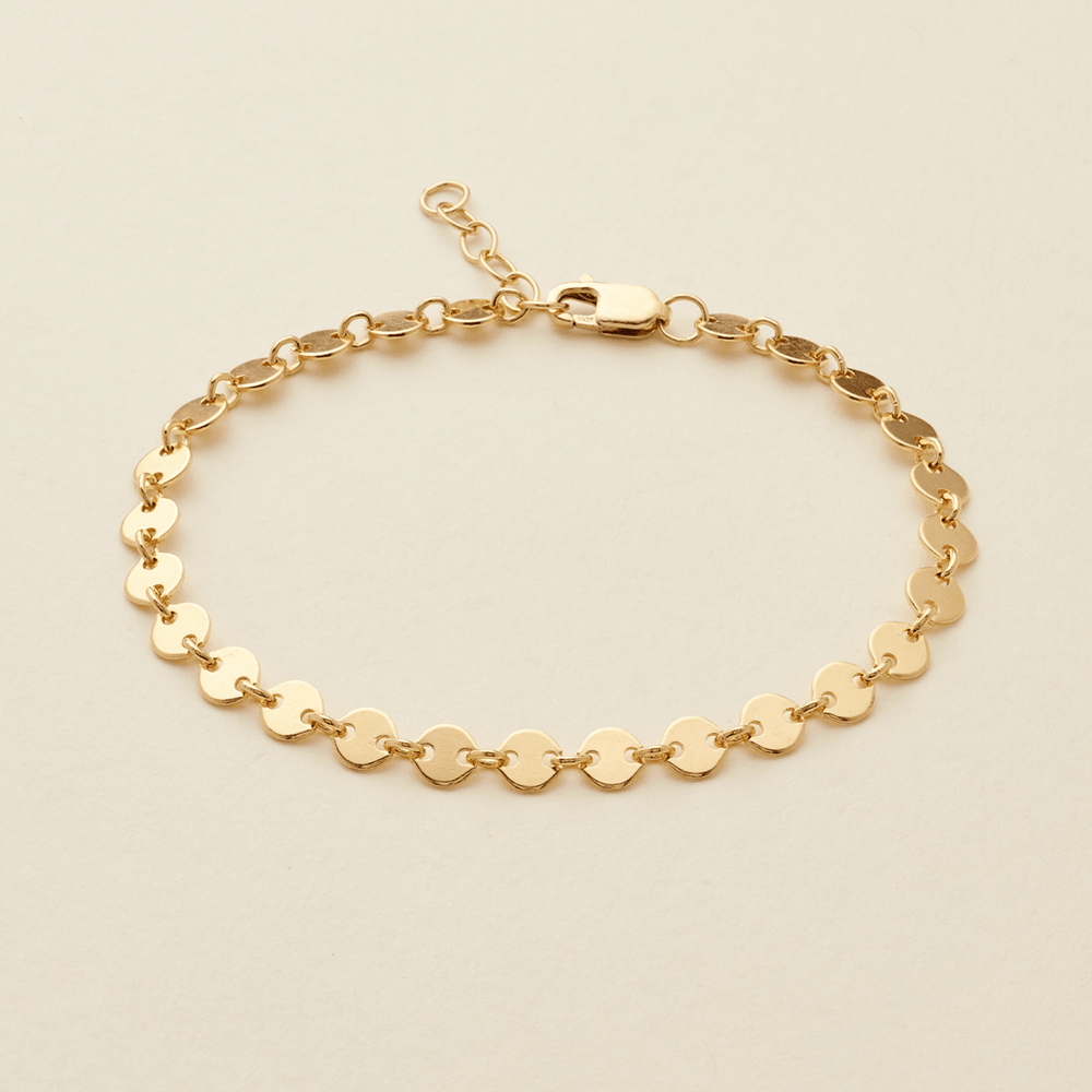 A gold chain bracelet with flat, circular disc links, displayed on a light beige background.