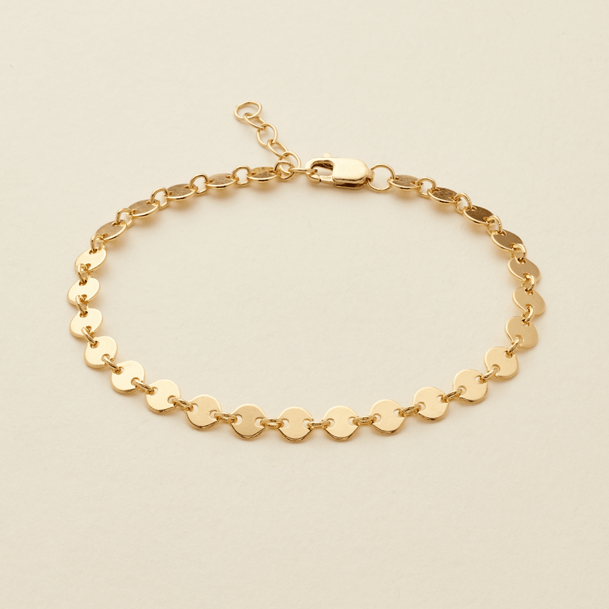 A gold chain bracelet with flat, circular disc links, displayed on a light beige background.