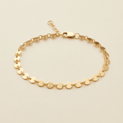 A gold chain bracelet with flat, circular disc links, displayed on a light beige background.