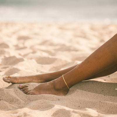 A close-up of a person's legs on the beach in sand showcasing a gold delicate chain anklet with round flat discs.