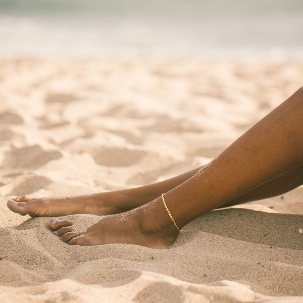 A close-up of a person's legs on the beach in sand showcasing a gold delicate chain anklet with round flat discs.
