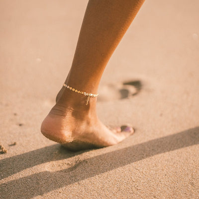 A close-up of a person's bare foot in sand showcasing a gold chain anklet with flat round discs linked together.