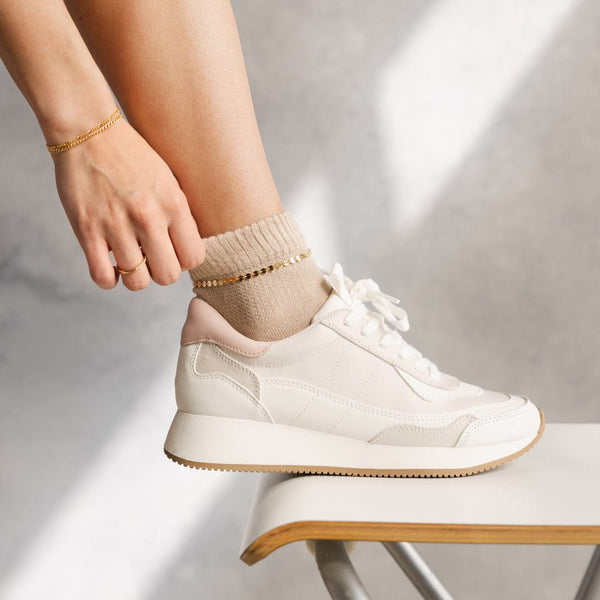 A close-up of a person's foot wearing white sneakers and beige socks, showcasing a gold anklet with flat round discs linked together, paired with matching gold bracelets and rings.