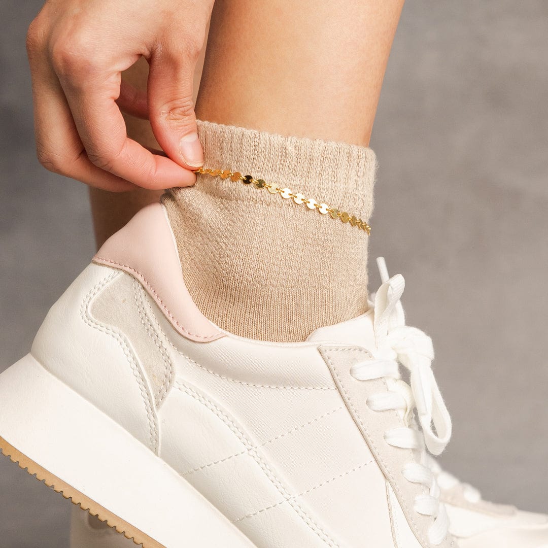 A close-up of a person's ankle wearing white sneakers and beige socks, showing off a gold delicate anklet made of round flat discs.