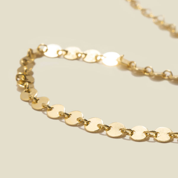 A close-up of a gold anklet made of flat round discs linked together, laying on a solid beige background.