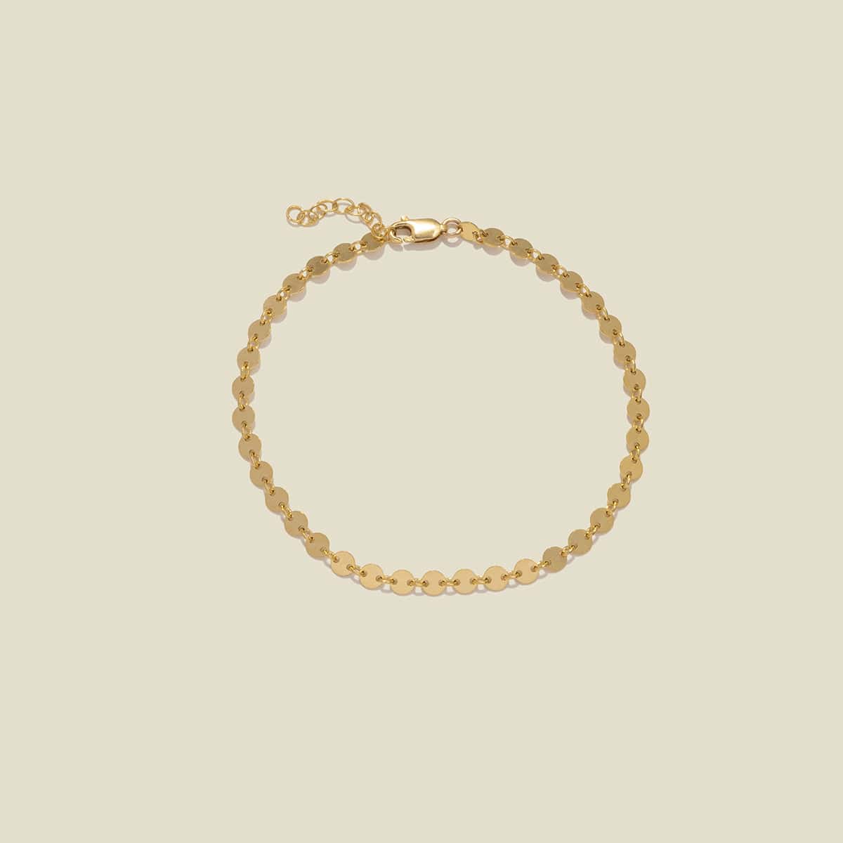 A delicate gold chain anklet with flat, circular disc links, displayed on a solid light beige background.