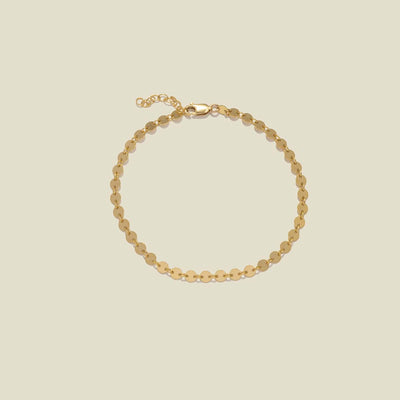 A delicate gold chain anklet with flat, circular disc links, displayed on a solid light beige background.