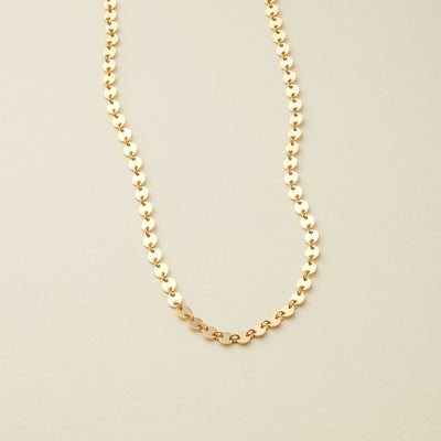 A delicate gold chain necklace with small, flat circular disc links, displayed on a light beige background.