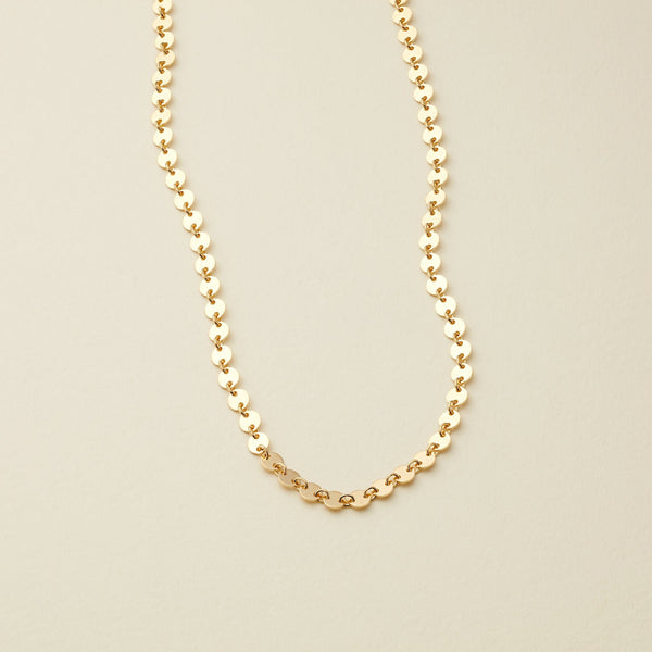 A delicate gold chain necklace with small, flat circular disc links, displayed on a light beige background.