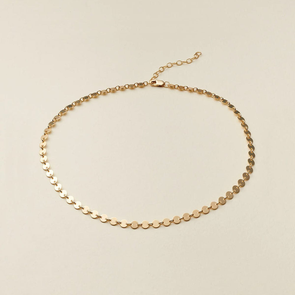 A delicate gold chain necklace with small, flat circular disc links, displayed on a light beige background.