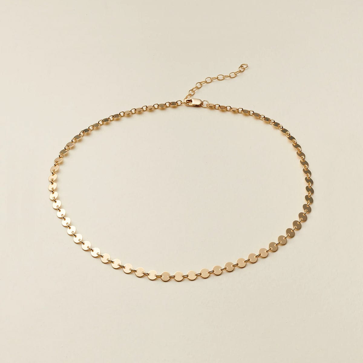 A delicate gold chain necklace with small, flat circular disc links, displayed on a light beige background.