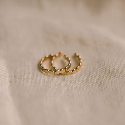  A pair of gold textured cuff earrings, laying on top of each other on a beige linen background.