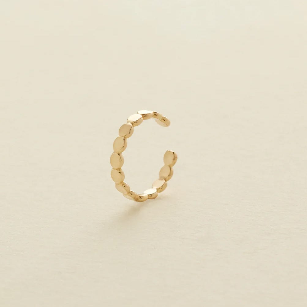 A delicate gold cuff earring made up of round flat discs laying side by side photographed on a clean, light beige background.
